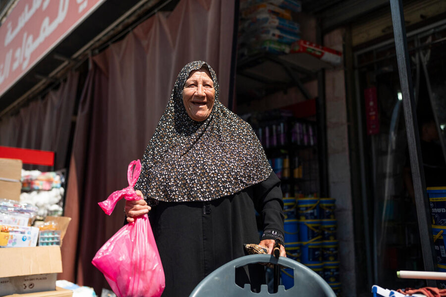 A smiling elderly woman in a dark gown and dark patterned headscarf holds a pink plastic bag as she emerges from a small shop. Photo: WFP/Hugh Rutherford