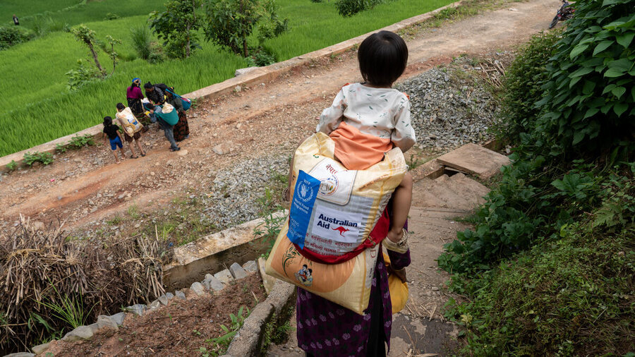 A women in a colorful outfit carries a bag of WFP food assistance and a child on her back down steep terrain in Nepal, with other people in the foreground. Photo: WFP/Shrawan Shrestha