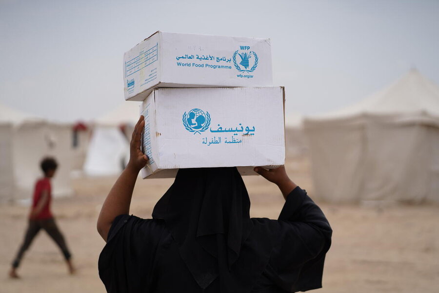 A person carries two WFP and UNICEF food boxes on their head in a camp setting