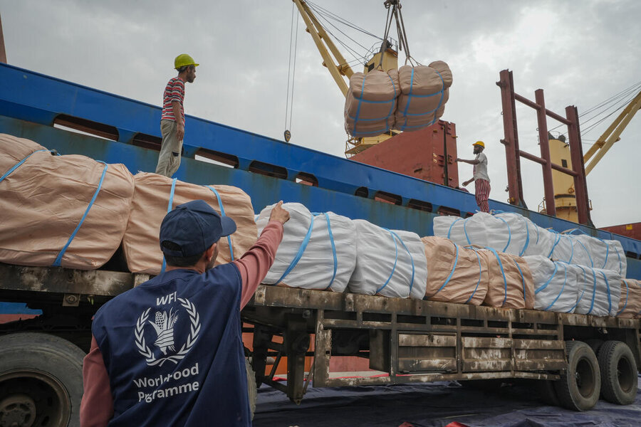 Workers load large cargo bundles between a ship and a truck at a port while a WFP staff member oversees operations