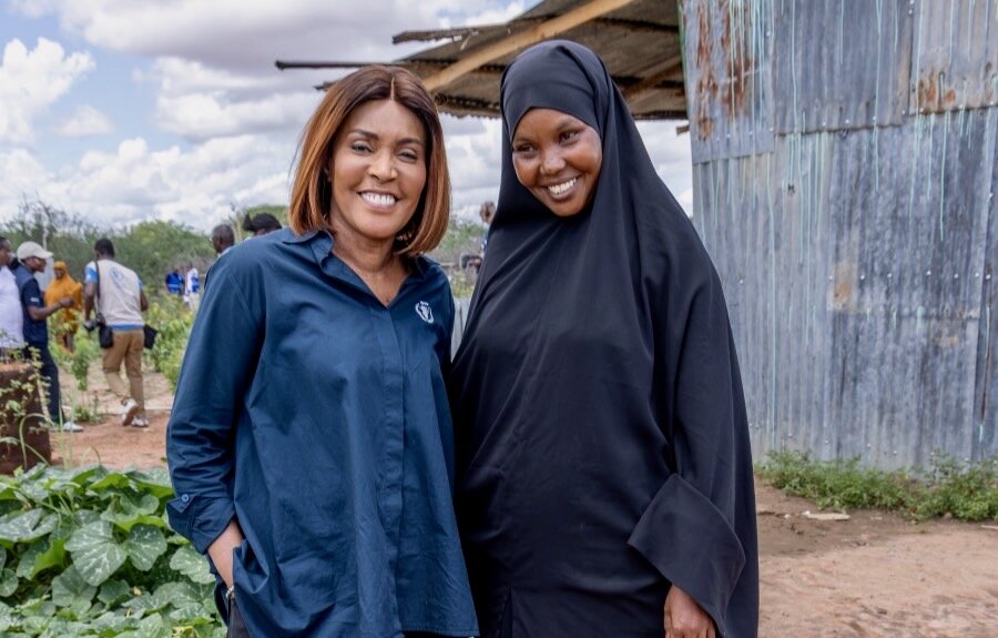 A woman in a long blue shirt and the other wearing a black hijab stand on the outskirts of a field, with people and a corrugated metal building behind them. hoto: WFP/William Orlale