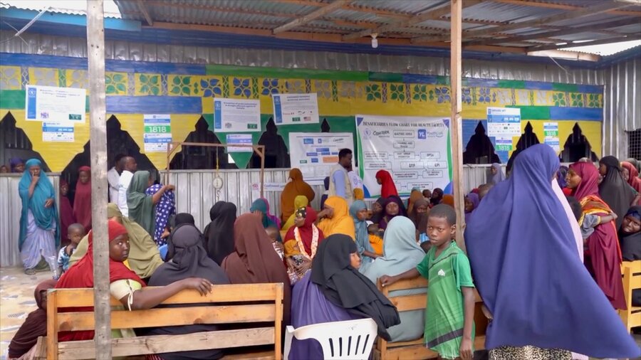 Children and colorful robes pack wooden benches and plastic chairs at a Mogadishu-area nutrition clinic, with colorful posters and a corrugated tin roof. Photo: WFP/Sylvie Njobati