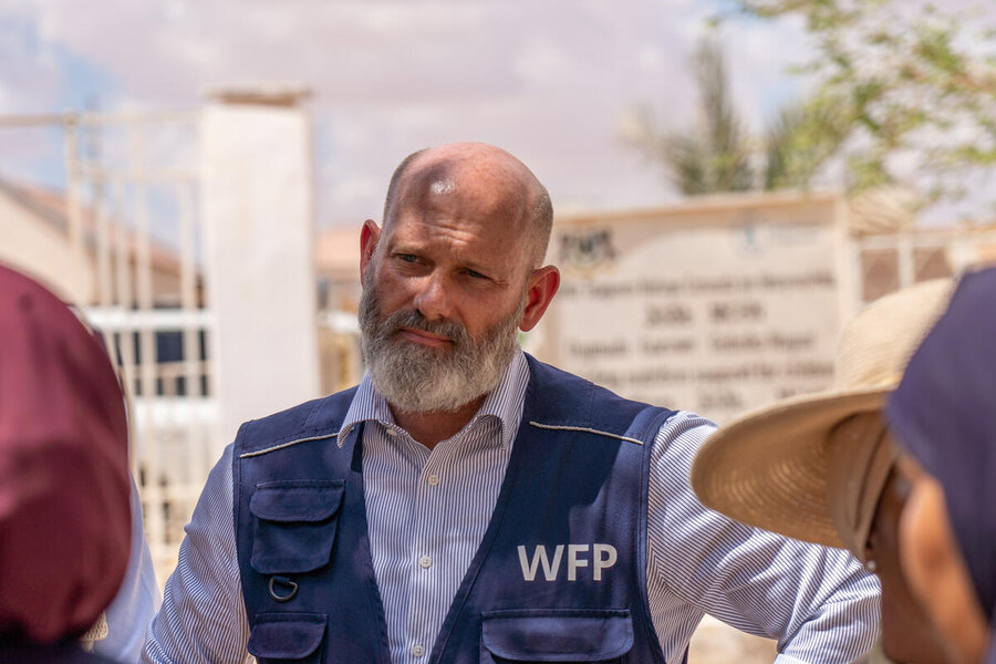 A WFP staff member wearing a blue vest speaks with people during a field visit, standing outdoors near aid facilities and community structures.