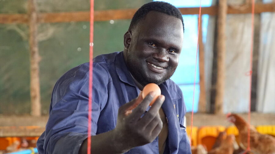 A smiling man in a blue smock holds up an egg, backdropped by chickens. Photo: WFP/Machrine Birungi