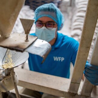Person in blue WFP shirt, hairnet and gloves operates machine dispensing grains into container.