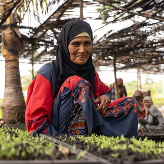 Person in black headscarf and red-blue patterned clothing sits on ground among small plants in shade, with other people in the background.