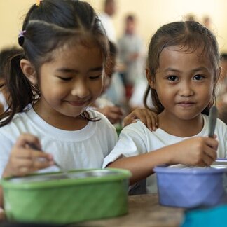 Two children in white shirts sit closely, eating from bowls with spoons.