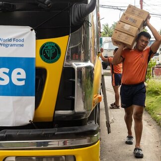 Person carrying three boxes on shoulder walks beside yellow truck displaying a World Food Programme banner.