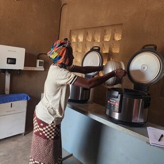Person in colourful attire uses a large electric cooker in a modest room with solar-powered units, highlighting sustainable cooking technology.