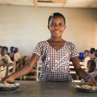 A young student smiles at the camera while holding onto two meals with either hand, her arms extended.