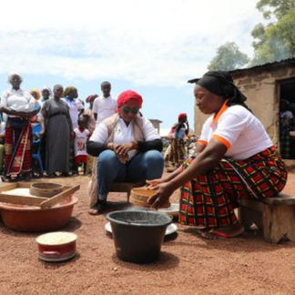 Women are seated and prepare food together while the community looks on