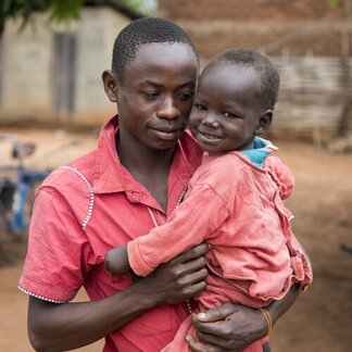 Person in red shirt holding child in matching red outfit, standing outdoors in a rural setting with visible structures and objects in background.