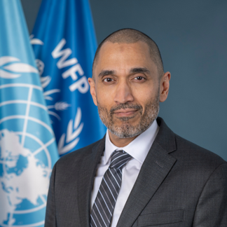 A portrait of WFP's Chief Financial Officer a.i. Ally-Raza Qureshi, standing in front of blue flags representing WFP and the United Nations
