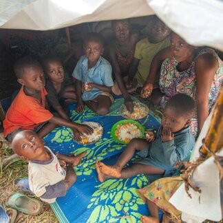 Children and an adult sit on a blue-green mat in a tent, sharing a meal from plates and a green bowl