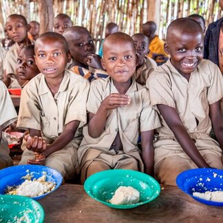 Children in school uniforms sit on a bench with plates of food in front of them, with other children in the background.