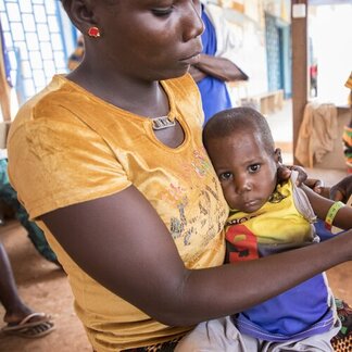 Person in yellow shirt holding child with colourful outfit and armband for checking malnutrition, in indoor setting with people in background.
