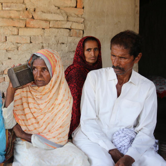 Three people gather outside a home with exposed brick walls. One person listens to a portable radio while others sit nearby, suggesting access to information or community communication.
