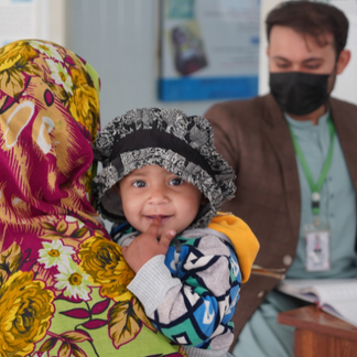 A young child looks toward the camera while being held by an adult wearing a colorful floral shawl. A person sits in the background inside a health or nutrition facility.