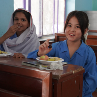 Two schoolchildren in a classroom eat a nutritious snack at their desks. A bowl containing fruit sits in the foreground while classroom benches and windows are visible in the background.