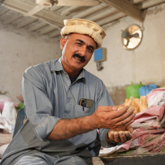 A person sits indoors holding grains in both hands. Sacks of agricultural products are stacked in the background of a storage or processing facility.