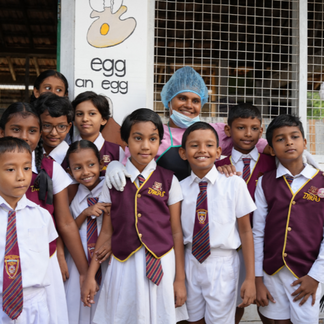 A group of schoolchildren in uniform standing together with a staff member outside a school building decorated with an egg-themed mural.