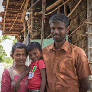 A family of a man, woman and child stand together outside in front of a wooden structure.