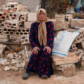 A person sits on a low stack of rubble and concrete blocks in a damaged outdoor area. The person wears a long patterned garment and a headscarf. Broken cinder blocks, debris, and a sack are arranged behind and beside the seated figure.