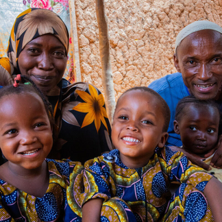 A smiling family of children sit together outside a mud-brick building, wearing brightly patterned clothing.