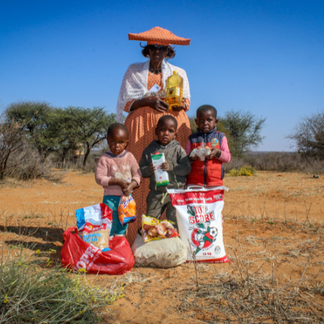 Caregiver stands with three children in a dry rural landscape, holding a bottle of cooking oil, while the children stand beside bags and cartons of food assistance including maize meal, pasta and other staples.