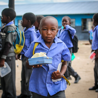 Child in a blue school uniform holds a container with a prepared meal while standing in a schoolyard, with other students and a school building in the background.