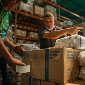 Workers organize and package food supplies in a warehouse, sealing and stacking boxes of fortified sunflower oil for distribution in Venezuela.