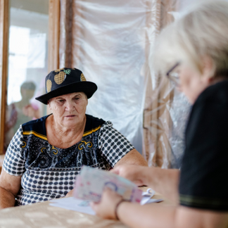 An older adult wearing a patterned hat sits at a table indoors while another person, partially out of focus in the foreground, counts or organizes banknotes. Plastic sheeting covers the wall behind them, suggesting a temporary or damaged setting, as cash assistance is being distributed.