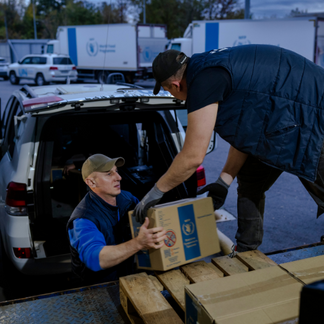 Two individuals transfer sealed cardboard boxes marked with the World Food Programme logo from the back of a vehicle onto a platform, with additional WFP trucks and vehicles visible in the background, illustrating logistics support and aid distribution.
