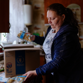 A person in a home kitchen removes packaged dry food items from a cardboard box labelled with the World Food Programme logo and places them on a counter, showing food assistance being received and organized indoors.