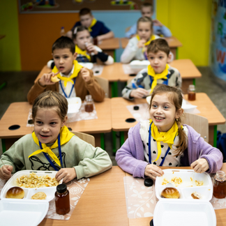 Children sit at desks in a brightly colored classroom, eating hot meals served in compartment trays with bread and drinks. More children are seated at desks in the background, illustrating a school meals setting.