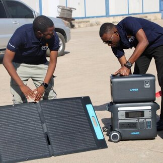 Two people in blue shirts set up a portable solar panel and power station outdoors, with a car and building visible in the background.
