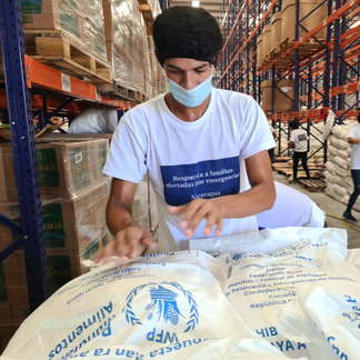 A colleague in a warehouse handling large white WFP food assistance bags, surrounded by stacked boxes and shelves of supplies