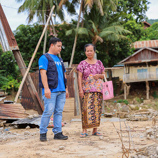 Two people stand on a dirt road in a village affected by flooding. Damaged structures and debris are visible nearby, with homes and palm trees in the background. The image shows an assessment visit in a community impacted by a disaster.