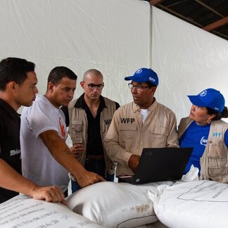 Group of people in WFP-branded clothing discuss logistics around large white sacks while viewing information on a laptop.