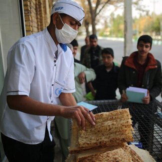 A WFP worker in white uniform and facemarks checks flatbread while people wait in line behind.