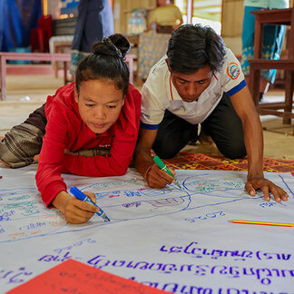 **Alt text:**   Two participants kneel on the floor and draw on a large sheet of paper during a community workshop. Colorful markers and handwritten notes are visible on the paper, while tables and chairs can be seen in the background of the indoor meeting space.