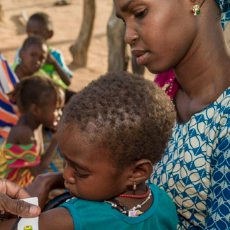 Caregiver holds a young child during a community nutrition activity, with other children gathered in the background outdoors.
