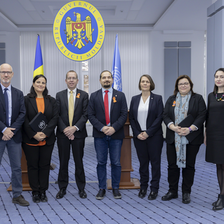 A group of seven people standing in a row, in front of the Moldovan government emblem and flags. They are dressed formally.