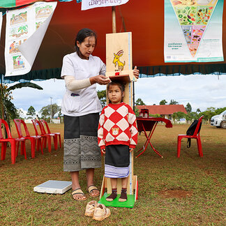 A child stands on a height measurement board outdoors while a healthcare worker records the measurement. Nutrition education posters hang inside a tented area in the background, with chairs and weighing equipment set up on the grass as part of a community nutrition screening activity.