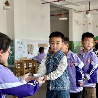 A young child holds a bowl while receiving food from a caregiver wearing gloves in a bright classroom setting. Several other children stand in line nearby, and educational posters and classroom furniture are visible in the background, illustrating a school-based nutrition activity.