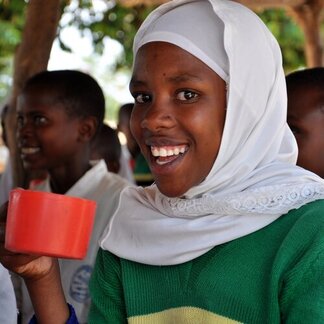 A young girl in white headscarf and green sweater holding a red cup.