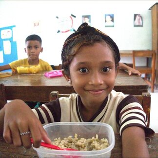 A child in a classroom sits in front of a food container, holding a spoon and ready to eat. Other children sit at desks in the background with posters and decorations on the walls.