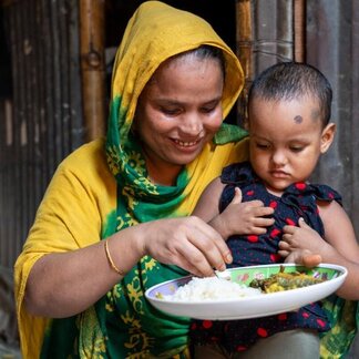 Bangladeshi mother in yellow dress and headscarf smiles as she feed her toddler from a plate