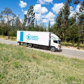 A large World Food Programme (WFP) delivery truck drives along a rural road bordered by grass and tall trees under a bright blue sky with scattered clouds