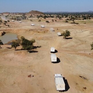 Convoy of trucks with white covers drives on a dusty road through an arid landscape with sparse trees and a small water body nearby.